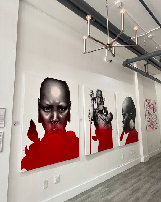 An art gallery interior displaying three large canvas portraits featuring Black subjects with striking red color accents on their clothing, hung beneath a modern geometric chandelier.