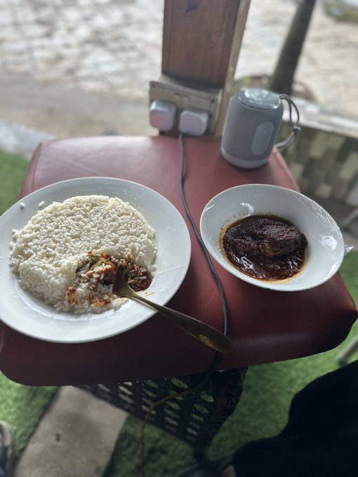 A meal of white rice and dark stew served on white plates on a red stool, with a mug and electrical outlet visible in the background.
