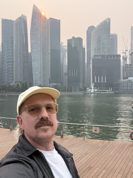 A man with a mustache, yellow cap, and tinted sunglasses takes a selfie against a hazy cityscape featuring modern skyscrapers by a waterfront.