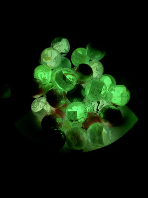 A close-up photograph of glowing green glass marbles illuminated in the dark, each containing various small illustrations and designs inside.