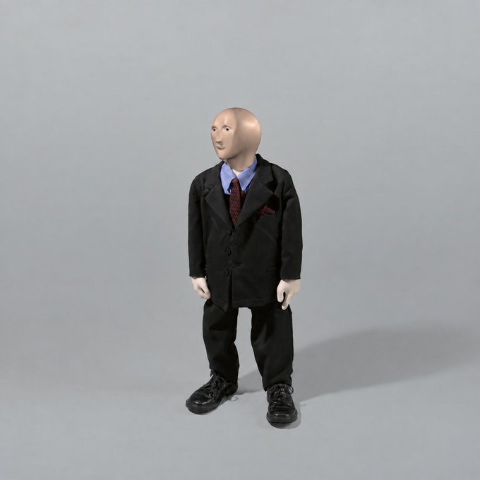A small figurine of a bald man wearing a black suit, blue shirt, and red tie stands against a plain gray background.