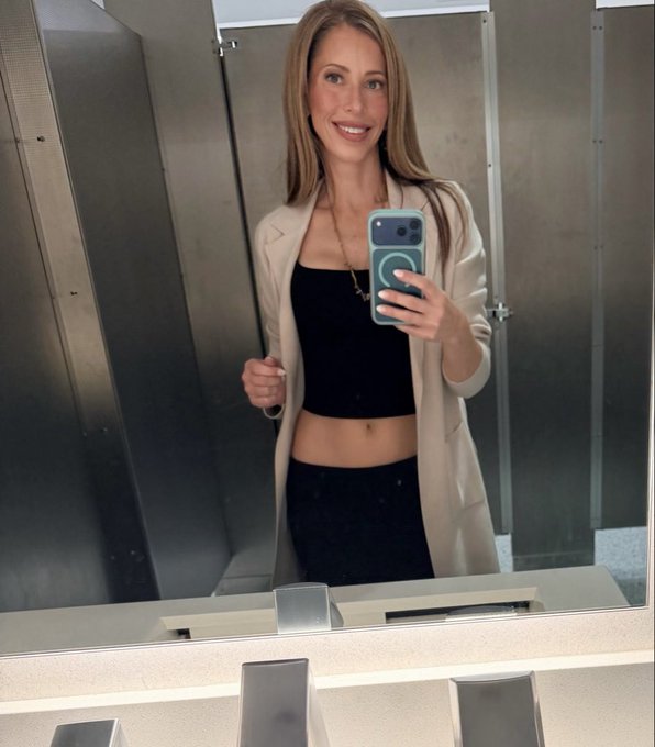 A woman taking a mirror selfie in what appears to be a public restroom, wearing a black crop top and cream-colored blazer while holding a phone with a teal case.