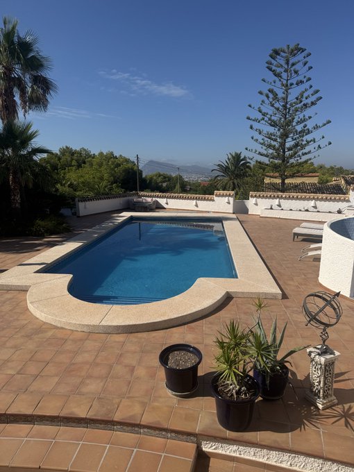 A serene Mediterranean villa with a tiled pool deck, potted plants, and a sun-drenched swimming pool overlooks rolling hills under a clear blue sky — a peaceful morning view fitting the cheerful "Herzlich guten morgen!" greeting.