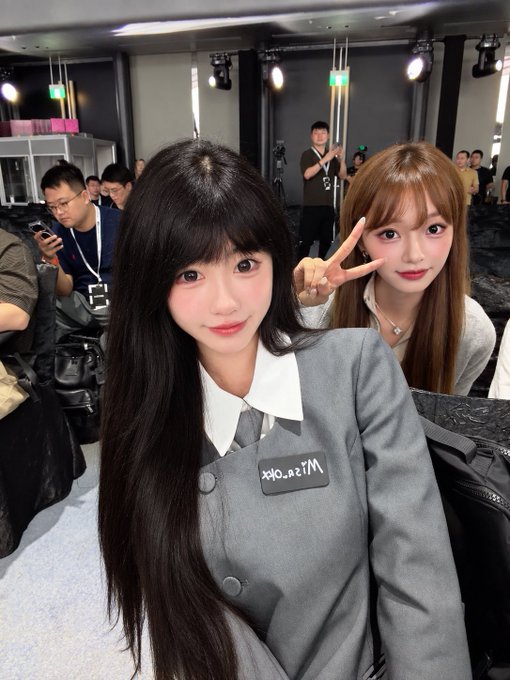 Two women pose for a selfie at what appears to be an event; the woman in front wears a gray uniform with a name tag reading 'Miss_fox' and the woman behind makes a peace sign.