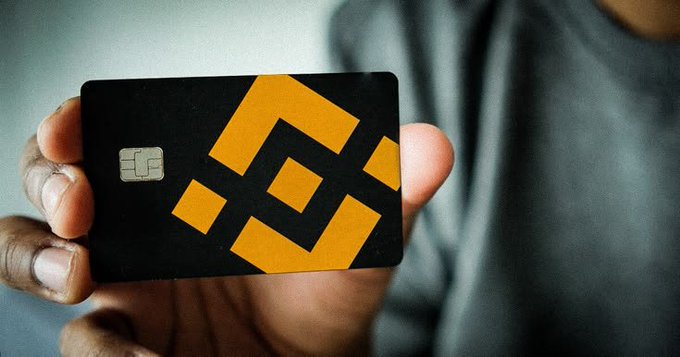 A close-up shows a person's hand holding a black Binance card featuring an orange geometric logo and an EMV chip.