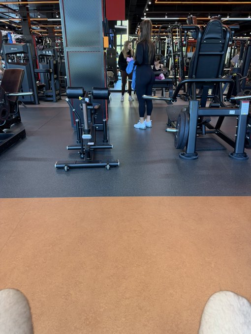 A view from someone sitting in a gym, showing their feet in the foreground while two women stand near exercise equipment in the background.