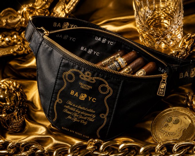 A premium product photograph of a black 'BAYC' (Bored Ape Yacht Club) fanny pack with gold lettering, paired with cigars, displayed on golden silk fabric alongside gold chains and a BAYC coin.