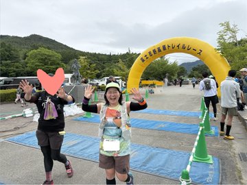 The image features a woman holding up her arms with a heart shape, possibly celebrating or cheering for someone. She appears to be at an event where she has crossed the finish line of a race. The setting includes a yellow archway that marks the end of the race and a green tarp on the ground, which could indicate a temporary boundary or a designated area for participants. There are several other people in the scene as well, suggesting that this is a popular event with multiple participants.