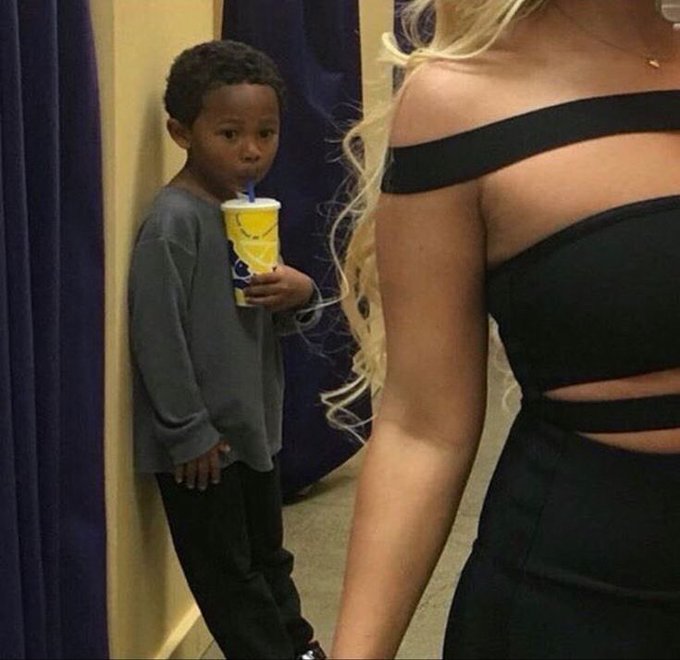 A viral internet meme photo showing a young boy holding a yellow cup, staring wide-eyed at a woman in a black dress standing nearby.