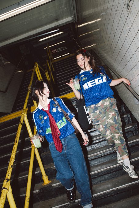Two young women pose on a stairwell in matching blue jerseys, with one displaying "MEGA" in bold white lettering across the chest—the apparent inspiration for the request to be seen in a @megaeth jersey. Their streetwear styling with camo pants and chain accessories captures the urban, on-trend aesthetic that responds to the post.