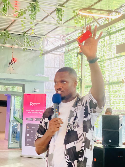 A man in a patterned shirt holds a microphone and gestures with a red card during a presentation, with a 'Rubiexclub Crypto' banner visible behind him.
