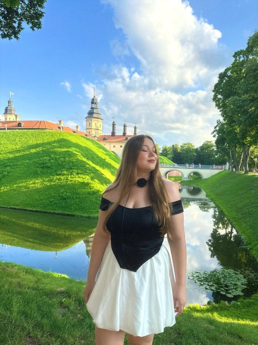 A woman in a black and white dress stands outdoors in a scenic European-style setting with green lawns, a pond, and a historic building with towers.