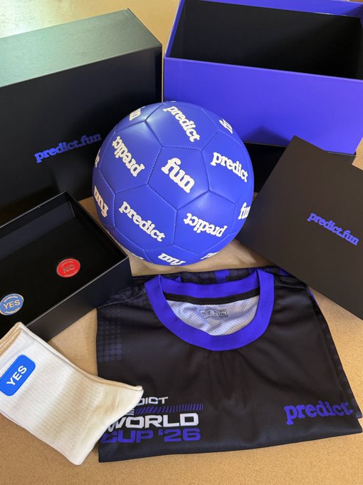 A promotional unboxing of predict.fun merchandise featuring a blue soccer ball branded with 'predict.fun' and 'fun' logos, a black 'World Cup '26' jersey, and accessories in branded boxes.
