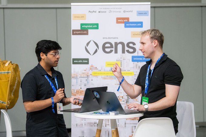 Two men wearing lanyards discuss at an ETHGlobal event beside a tall ENS Domains banner featuring the tagline "THE NAMES LAYER OF WEB3" and logos for PayPal, Coinbase, Uniswap, and Worldcoin, alongside various .eth addresses like "james.eth" and "wulfswap.eth." The scene illustrates the post's message about ENS being a partner sponsor offering $5,000 in prizes for using ENS as identity, discovery, and profiles for agents and wallets.