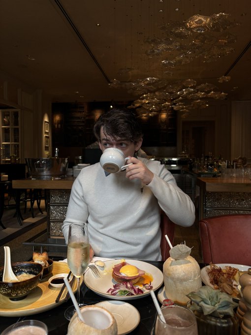 A young man sitting at a dimly lit restaurant table taking a sip from a cup, surrounded by various plated dishes, drinks in coconuts, and champagne.