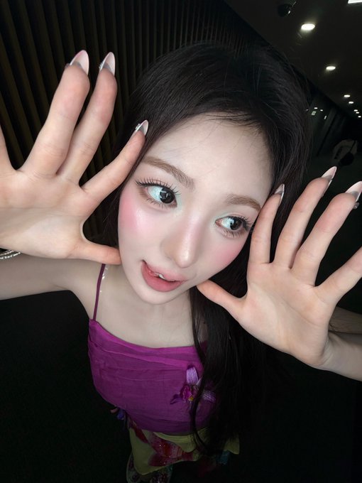 A young woman with long dark hair poses playfully with both hands framing her face, wearing a purple floral-patterned top with spaghetti straps and long white manicured nails. The caption "Whine #KiiiKiii #키키 #SUI #수이" suggests this is a selfie of SUI from the K-pop group KiiiKiii, with hashtags in both English and Korean identifying her stage name and group.
