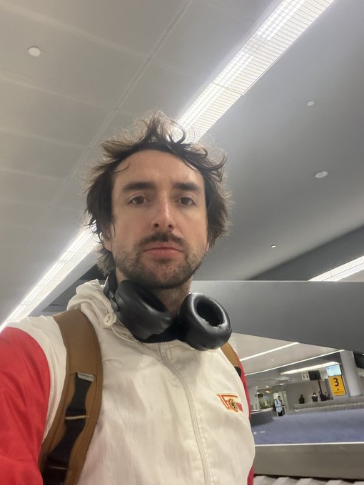 A man with messy hair and headphones around his neck takes a selfie in what appears to be an airport baggage claim area.