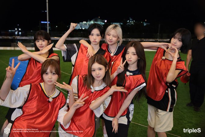 Eight young women in red sports jerseys pose playfully on a green field at night, with the 'tripleS' logo in the corner and a copyright notice for '2025 Modhaus All Rights Reserved.'