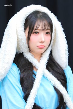 A close-up portrait of a young woman with long black hair wearing a light blue top and a large, fluffy white hood with ear flaps.