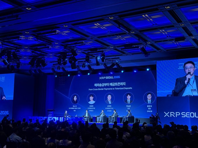 A panel at XRP Seoul 2026 with the title "From Cross-Border Payments to Tokenized Deposits" features five seated speakers pictured on screen, while a man with a microphone speaks at the right podium, as the audience watches the discussion on stage.