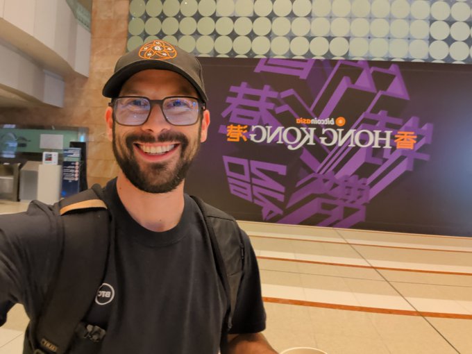 A man wearing glasses, a black t-shirt, and a cap smiles for a selfie at what appears to be a Hong Kong airport, with a "Discover Hong Kong" banner (featuring the text in both English and Chinese characters 香港) visible behind him—capturing the excitement of starting a new journey on Day 1.