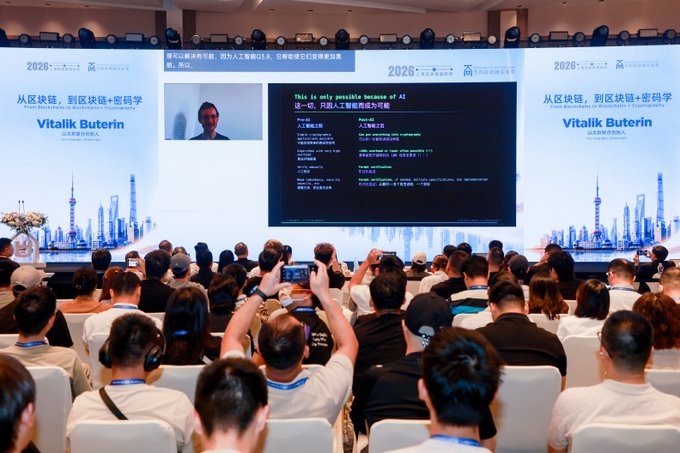 A conference audience watching a presentation on a large screen by Vitalik Buterin, with slides titled '从区块链，到区块链+密码学' (From Blockchain to Blockchain + Cryptography) and code snippets.
