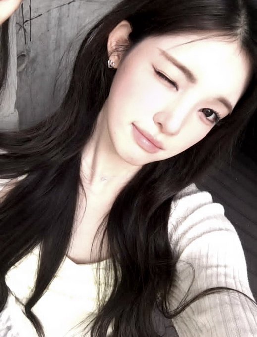 A close-up selfie of a woman with long dark hair winking at the camera, wearing a white ribbed top and earrings.