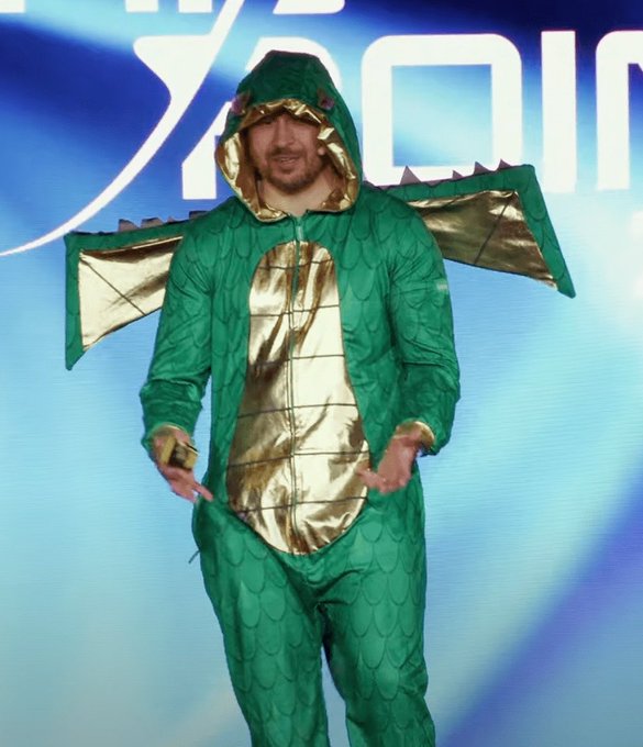 A man wears a green dragon costume with gold accents and wings, standing on stage in front of a partial "X" and "DIN" logo backdrop, likely referring to Vitalik Buterin of Ethereum. The costume visually represents a "Vitalik dragon," poking fun at the Solana founder's challenge in the accompanying post comparing the two blockchain creators.
