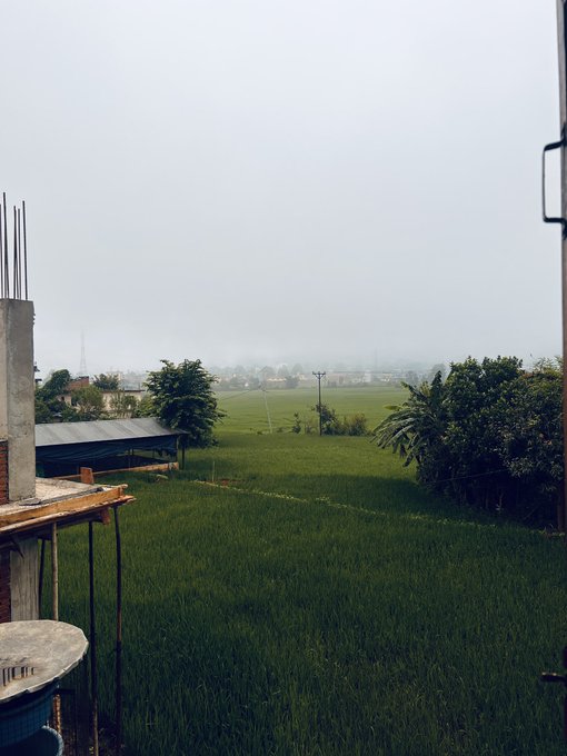 A view from a balcony overlooking lush green rice fields and distant village rooftops shrouded in misty, overcast weather, with a partially constructed building and scaffolding visible in the foreground.