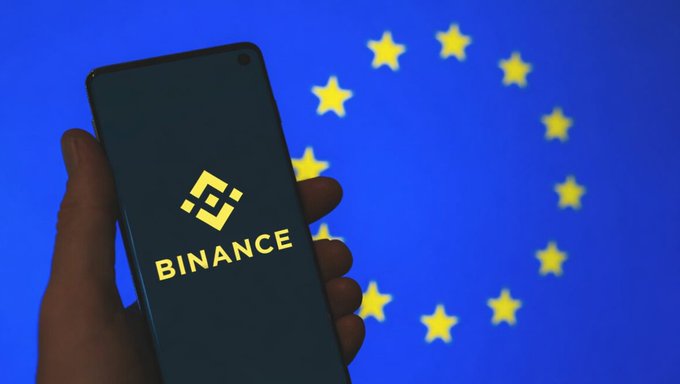 The image shows a person holding up their phone, which displays the Bitcoin logo. The background of the image features a blue circle filled with yellow stars, creating an interesting visual contrast. Below the image, there is text that reads "An Update on #Binance’s MiCA Regulatory Journey & Europe's Digital Asset Future There’s been a lot of uncertainty around MiCA lately." This suggests that the person holding up their phone might be sharing some information or news related to Bitcoin and its regulatory status.