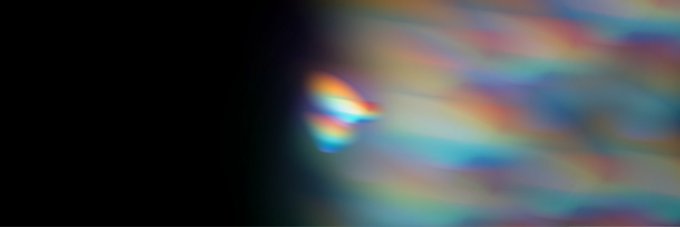 An abstract blurry image showing streaks of rainbow-colored light against a black background, resembling light leak or chromatic aberration effects.