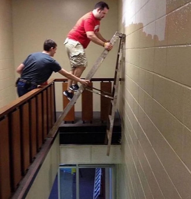 The image humorously captures the essence of the caption by showing two men precariously maneuvering an A-frame ladder on a narrow upper-floor balcony—one man balancing on the railing while the other guides the ladder over the edge, suggesting a daring, risky maneuver. There is no visible text, numbers, or tickers in the image, but the scene visually symbolizes the extreme and reckless risk-taking associated with memecoin traders.