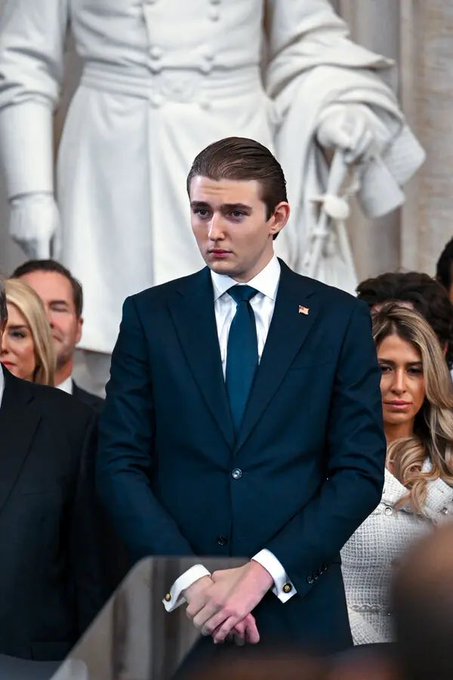 A young man with dark hair, wearing a dark blue suit and tie with an American flag pin, stands solemnly with his hands clasped in front of him.