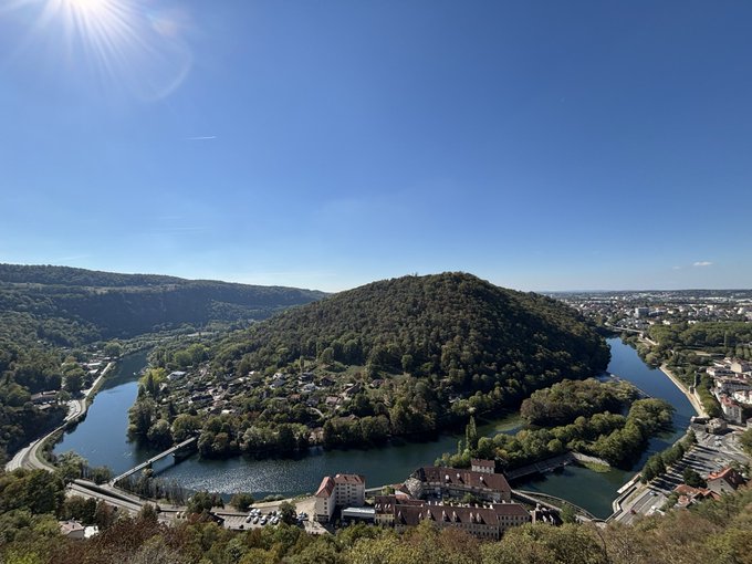 An aerial landscape of a town nestled along a winding river that forms a peninsula, with a forested hill in the center under a clear blue sky.