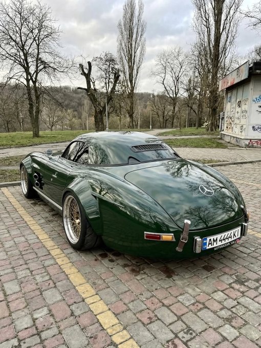 The image features a green sports car parked on a brick road, with a brick wall visible behind it. The car appears to be an antique Mercedes-Benz SLS-Class Coupe, which has been modified for racing purposes. The car is parked in front of a building and there are trees nearby as well.