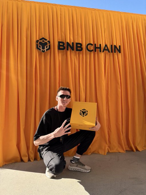 A man in a black shirt and sunglasses crouching in front of an orange 'BNB CHAIN' backdrop with a yellow branded cube.