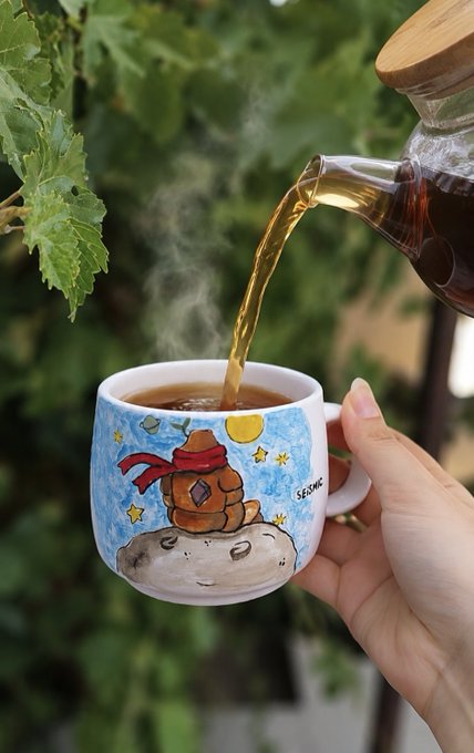 Hot tea pours from a glass pot into a hand-painted ceramic mug featuring a "Seismic" logo and an illustration of a small character sitting on a rock beneath stars and a sun, set against a lush green leafy background. The image captures a cozy morning ritual that visually represents the post's message about fresh starts and brewing new ideas for the week ahead with Seismic.