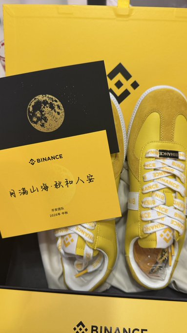 A pair of yellow and white Binance-branded sneakers displayed with a card featuring a moon graphic and Chinese text '月满山海·积和人生'.