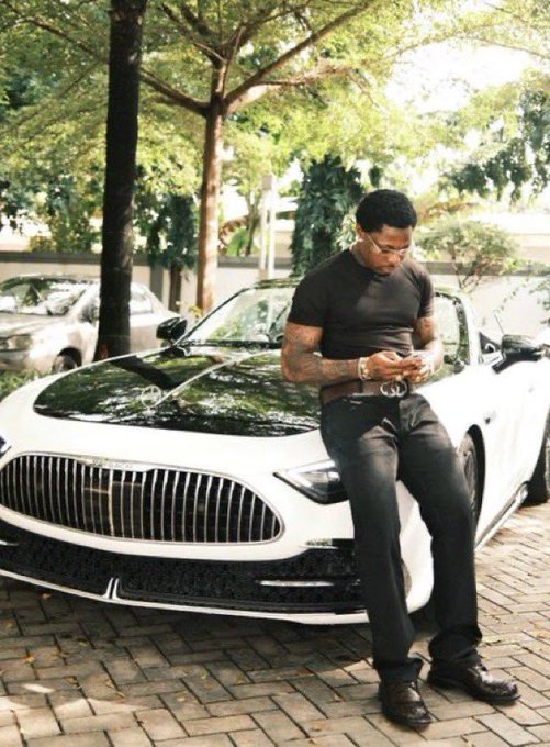 A man in a dark t-shirt and sunglasses stands leaning against a white and black Mercedes-Maybach luxury vehicle outdoors.