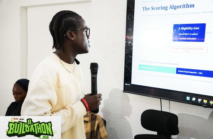 A young man with braided hair and glasses stands holding a microphone in front of a screen titled "The Scoring Algorithm" with a "2. Eligibility Gate" box visible, while a "Build-A-Thon 1.0 JOS" logo appears in the corner.