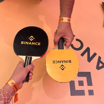 Two hands exchange branded Binance table tennis paddles at the Binance 9th Anniversary meetup, with the orange backdrop displaying the "BINANCE" logo and name. The image captures a fun, interactive moment that aligns with the post celebrating the community event and personal milestone of 6 years with Binance.