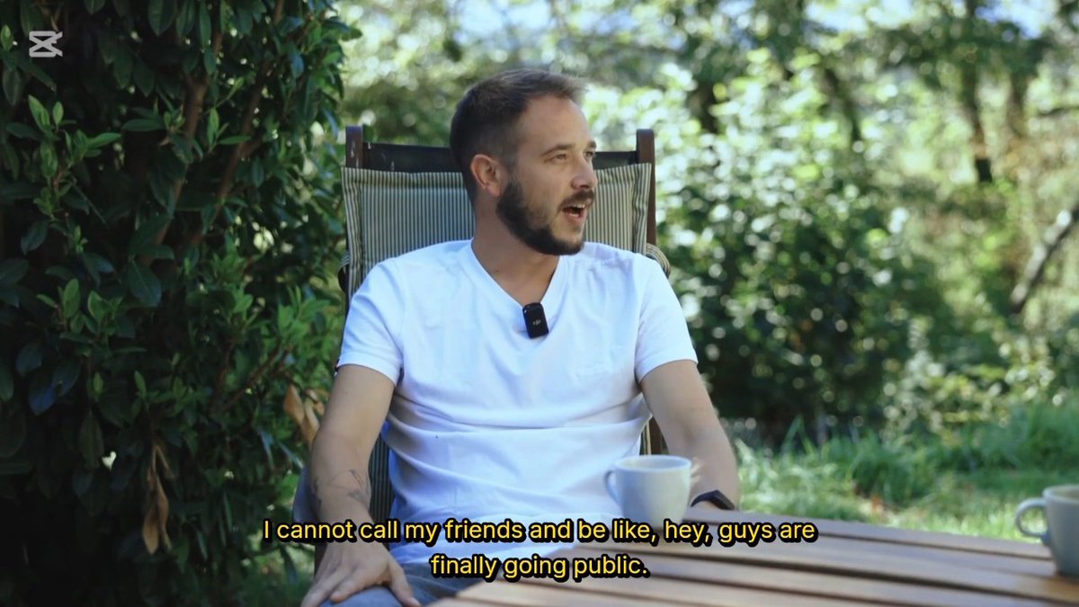 A man in a white t-shirt sitting outdoors with a microphone, with subtitles reading 'I cannot call my friends and be like, hey, guys are finally going public.'