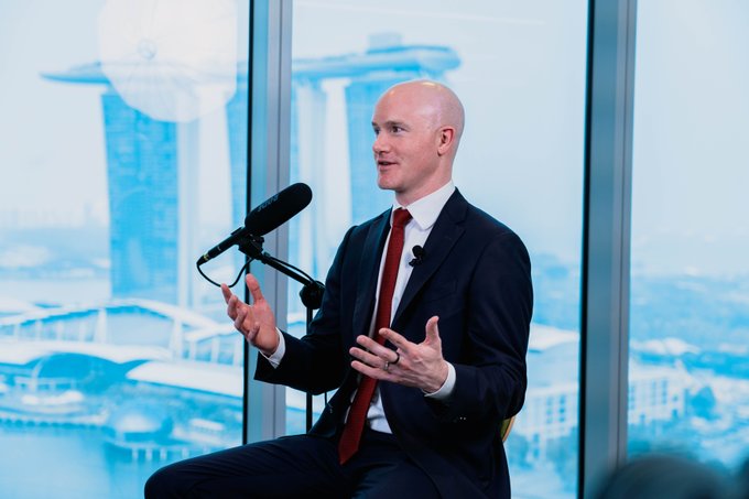 A bald man in a dark suit and red tie speaks while gesturing, seated in front of a microphone with the Marina Bay Sands hotel visible through the window behind him.