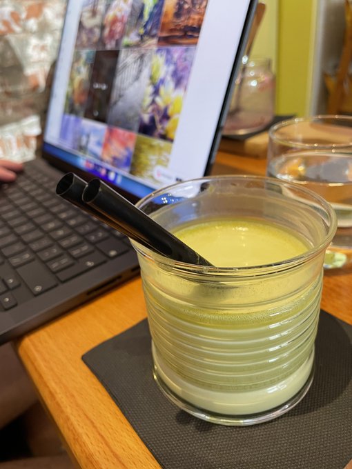 A close-up of a glass of green smoothie with a black straw on a coaster, with a laptop displaying an image gallery visible in the background.