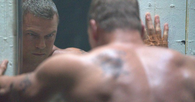 A shirtless, tattooed man with a buzzcut stares intensely at his reflection in a mirror, pressing his hands against the wall. The image evokes the gritty, hardened atmosphere of a revenge thriller, fitting the post about Alan Ritchson's "Motor City" and its commentary on Netflix's action movie conventions.