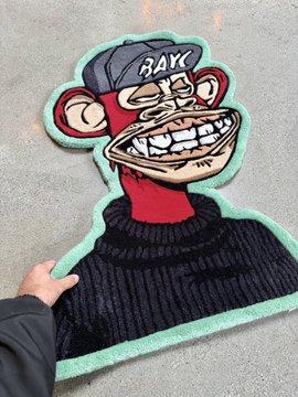 The image features a person holding up a rug with a cartoon monkey design, which includes a hat and a sweater. The rug has a green border around its edges, giving it an artistic touch. The caption "bayc otherside apecoin rugs Irl is alpha" suggests that the rug might be part of a collection or series by Bayc, a company known for their unique designs and illustrations.