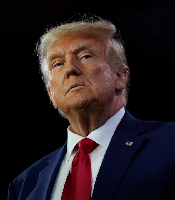 A close-up portrait of Donald Trump wearing a dark blue suit, white shirt, and red tie with an American flag lapel pin, set against a black background.
