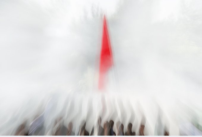 A blurry, motion-distorted image showing a red vertical shape in the center surrounded by white streaks radiating outward, with hints of a crowd at the bottom.