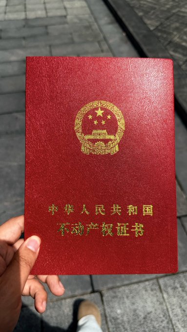 A first-person view shows a hand holding a red booklet with the gold national emblem of China, displaying the Chinese text '中华人民共和国 草命产饭证证书' (a garbled translation of 'People's Republic of China Property Ownership Certificate') over a paved ground background.