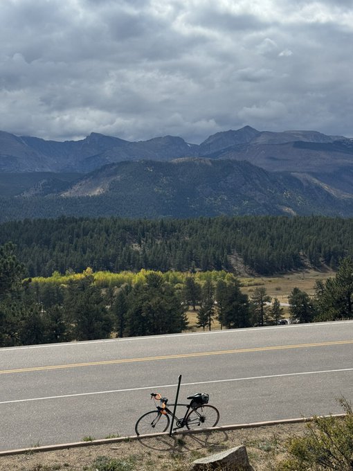 A bicycle with a rear pannier bag parked on the shoulder of a paved mountain road, with forested hills, rocky peaks, and a cloudy gray sky in the background.